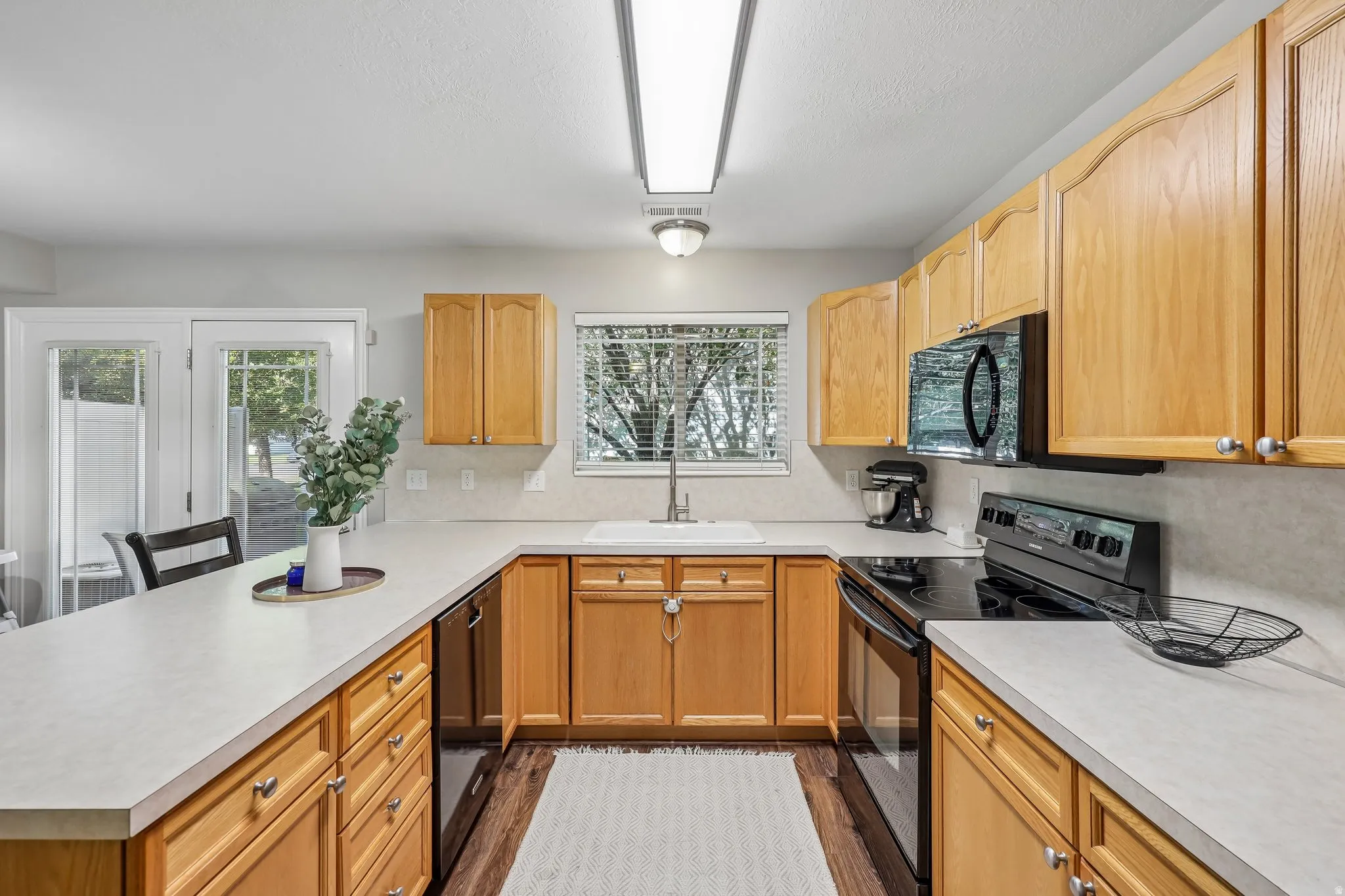 Kitchen featuring light wood cabinetry, light-toned countertops, a stainless steel sink, black appliances, and wood-finish flooring