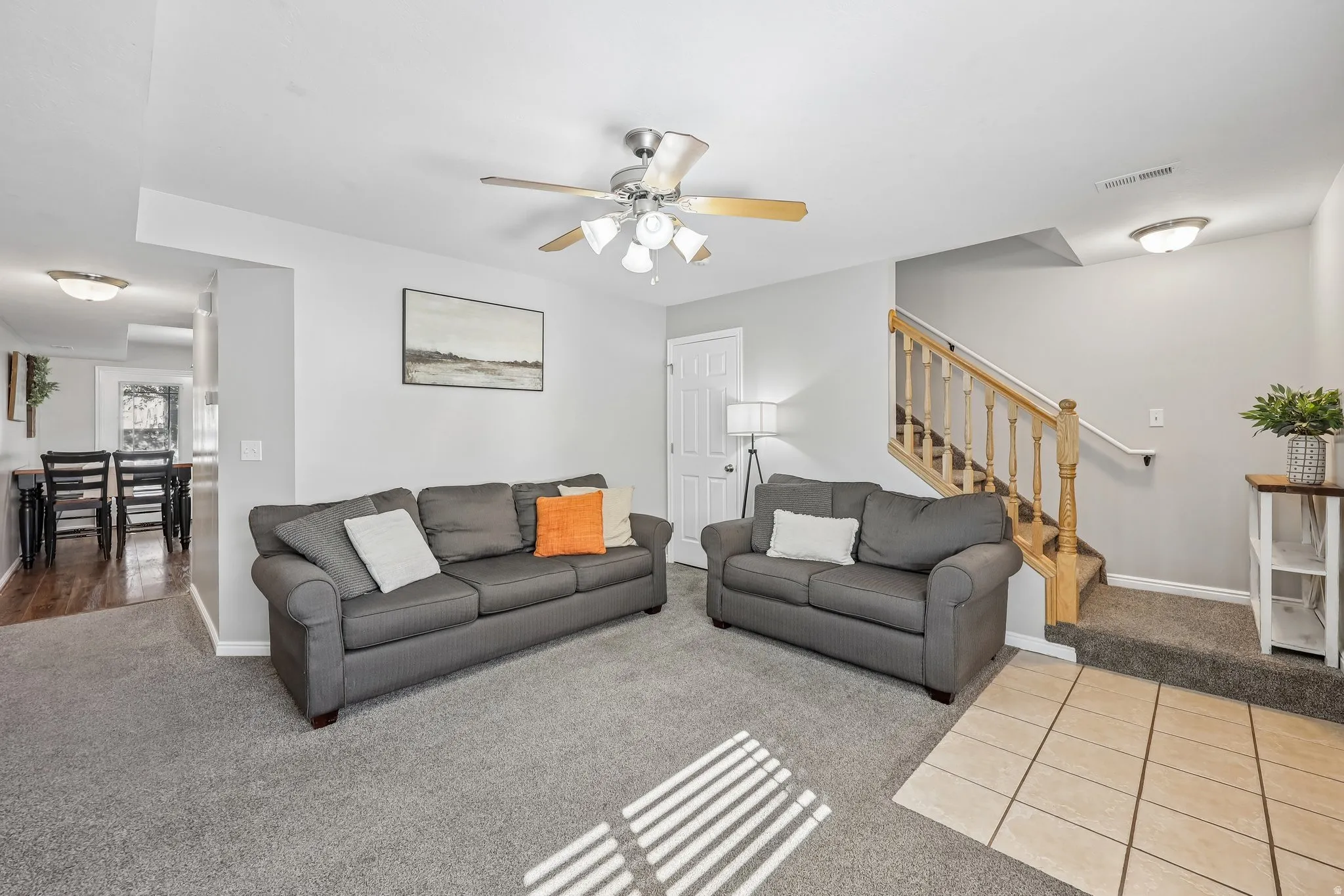 Carpeted living area with a ceiling fan light fixture, wood banister stairway, and tile flooring at the entry