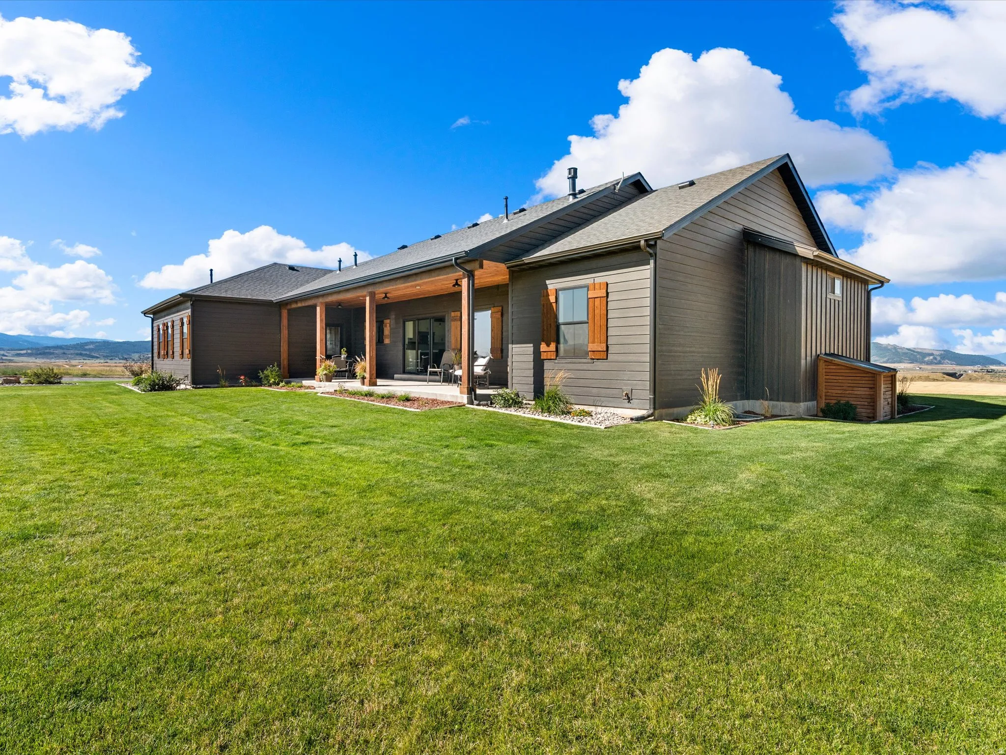 Expansive exterior featuring a covered patio with wood support beams, dark siding, and a vast lawn