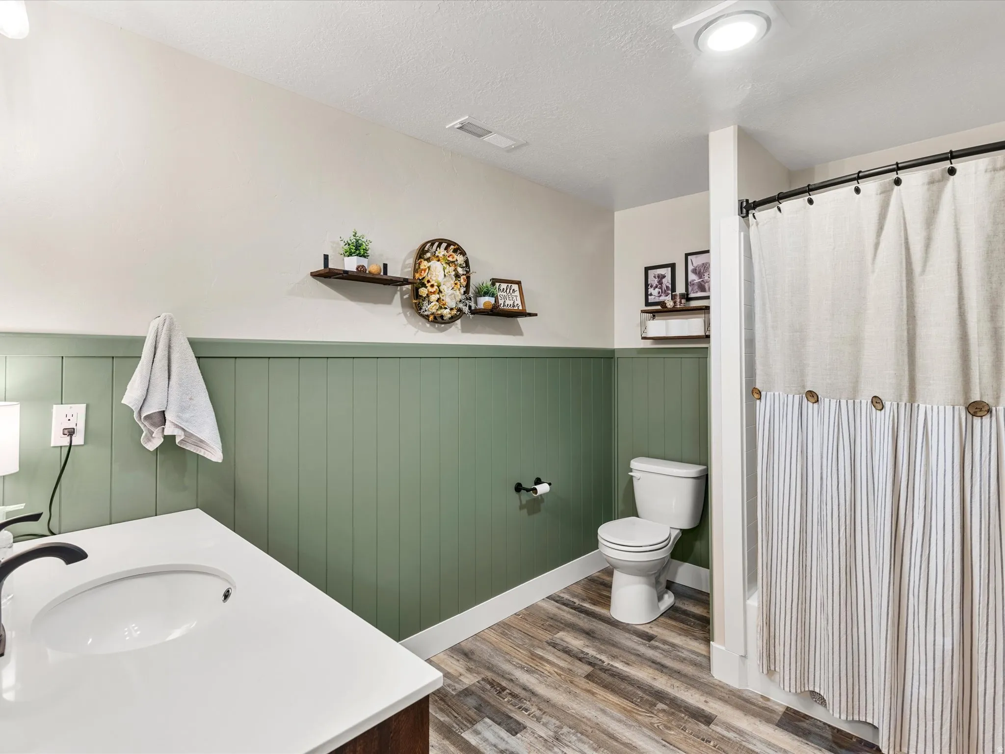 Bathroom featuring a white vanity with an integrated sink, wood-finish flooring, a toilet, and a shower with a curtain rod