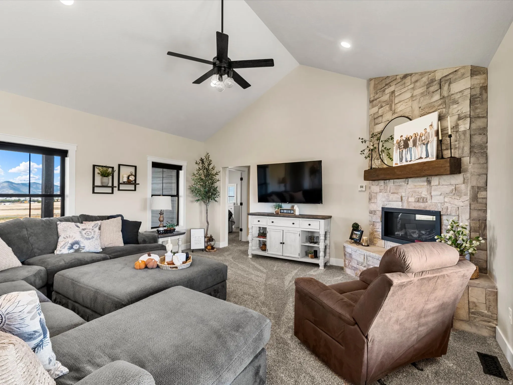 Vaulted ceiling with a ceiling fan, stone-clad fireplace with a wood mantle, large windows with black trim, and neutral carpet flooring