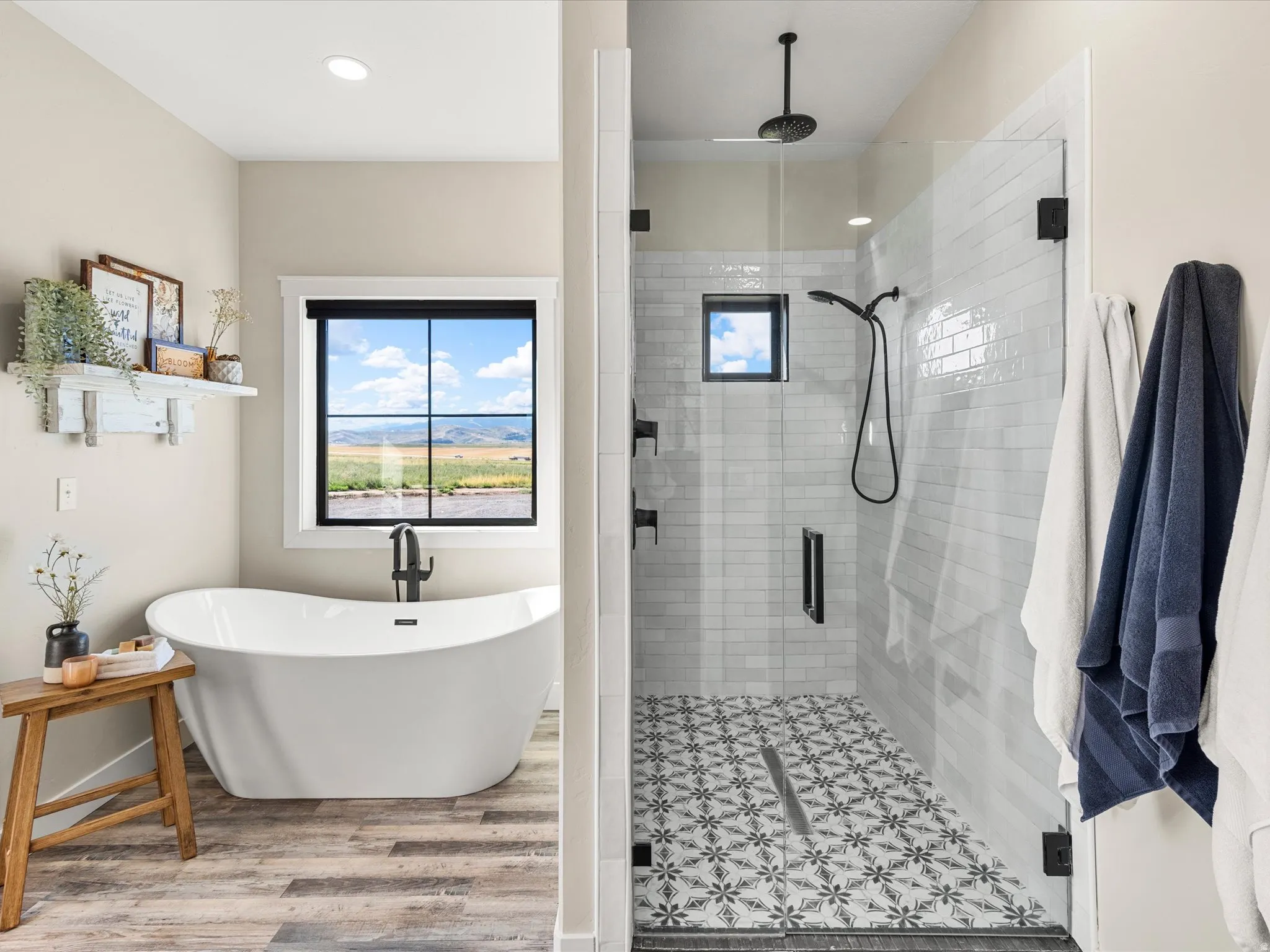 Freestanding soaking tub with wood-finish flooring, glass-enclosed shower with subway tile, patterned shower floor, and matte black fixtures