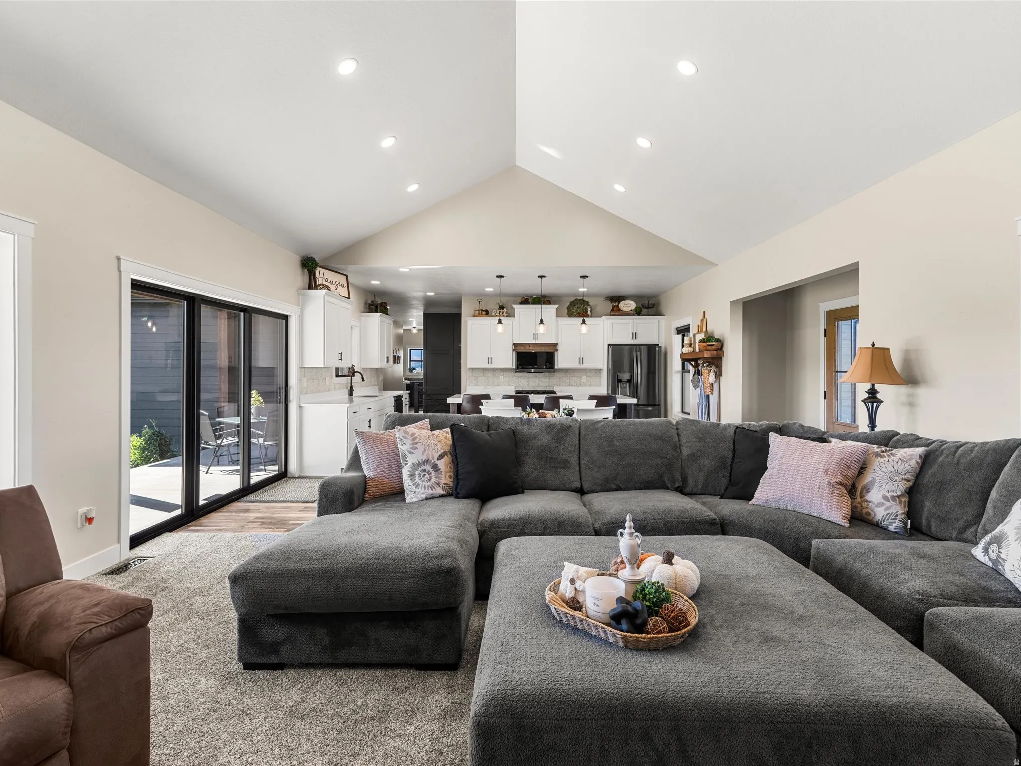 Vaulted ceiling with recessed lighting, black frame sliding glass doors, carpeted flooring, and a kitchen with white cabinetry and dark appliances