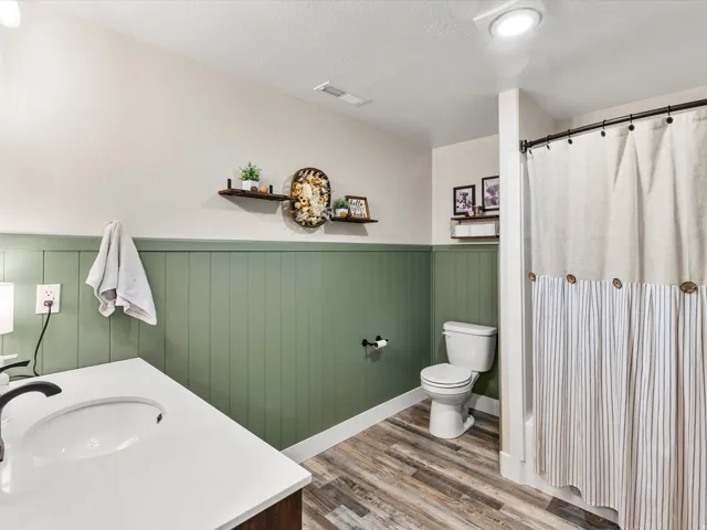 Bathroom featuring a white vanity with an integrated sink, wood-finish flooring, a toilet, and a shower with a curtain rod