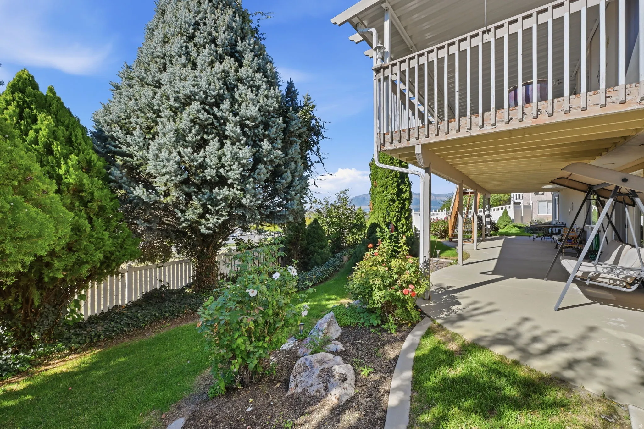 Lower level covered patio, upper deck with wood railing, established landscaping, and a white picket fence