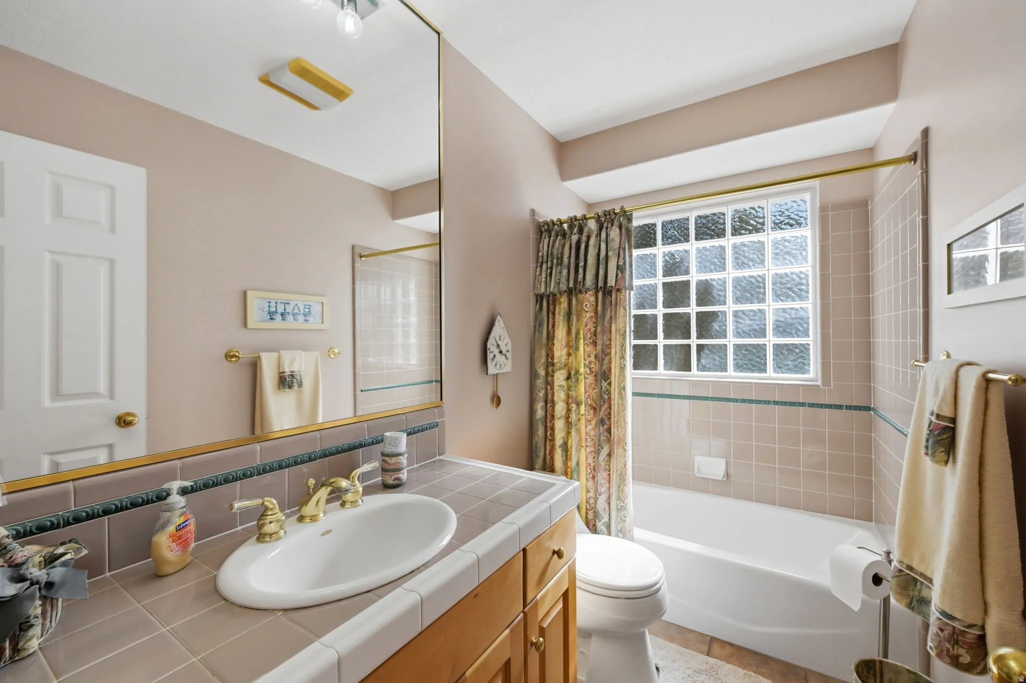Vanity with tiled countertop and integrated sink, full-width mirror, glass block window, built-in bathtub, and tiled shower surround