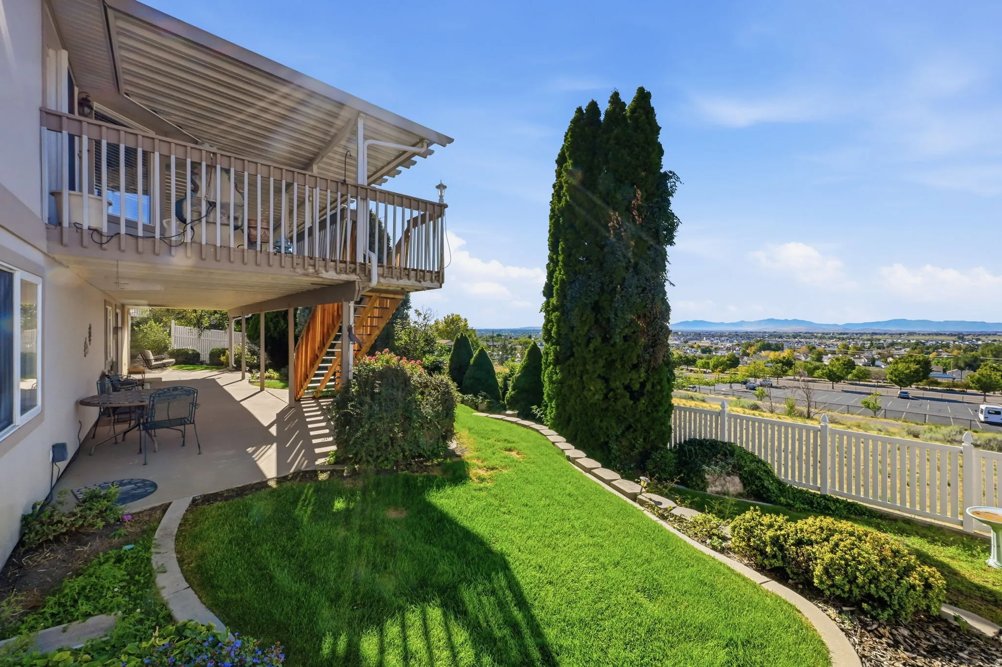 Expansive upper-level deck with wood railing, covered lower-level patio, sloped green lawn, and distant city scenery