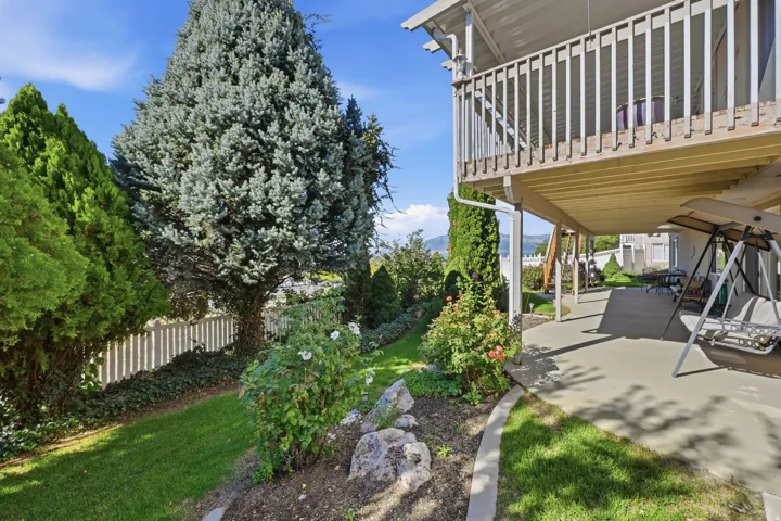 Lower level covered patio, upper deck with wood railing, established landscaping, and a white picket fence