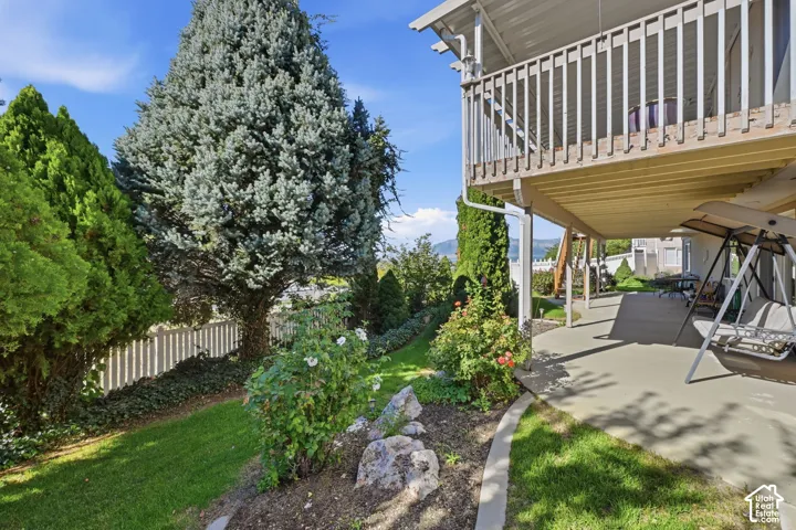Lower level covered patio, upper deck with wood railing, established landscaping, and a white picket fence