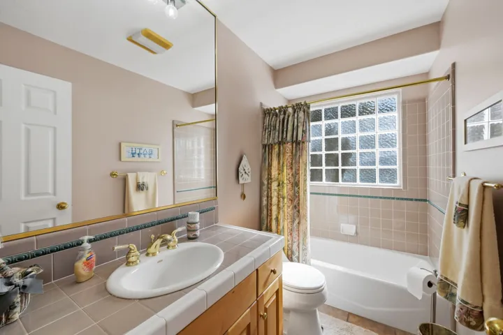 Vanity with tiled countertop and integrated sink, full-width mirror, glass block window, built-in bathtub, and tiled shower surround