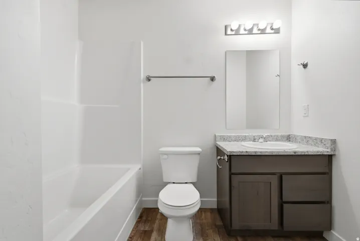 White bathtub and shower surround, wood-finish flooring, vanity with light-colored countertop, integrated sink, and dark wood-finish cabinetry
