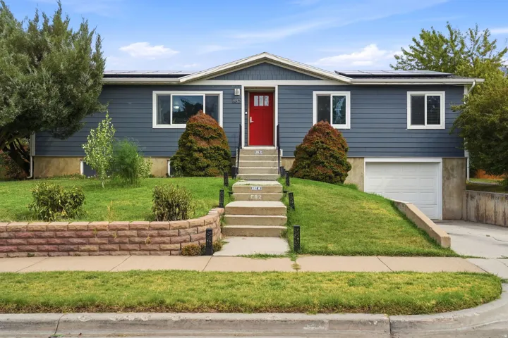 Two-story residence featuring a striking red front door, prominent solar panels, an integrated garage, and a retaining wall with landscaping