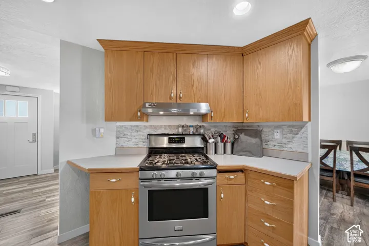 Stainless steel gas range with coordinating ventilation hood, wood-finish cabinetry, light-toned countertops, tiled backsplash, and recessed lighting