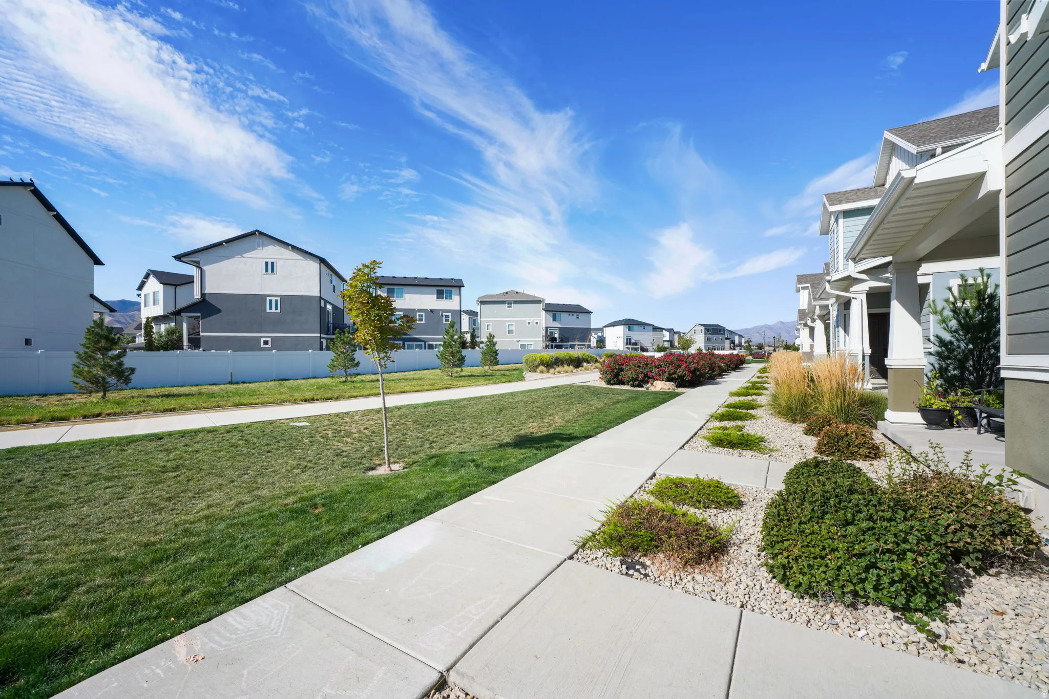 Landscaped walkway featuring decorative plantings, a green lawn, and a light-toned privacy fence