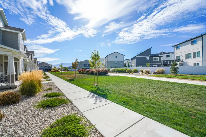 Exterior featuring a concrete walkway, established green lawn, mature landscaping, and a row of townhomes with covered porches
