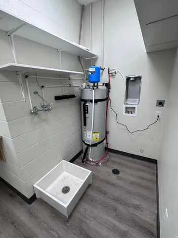 Utility area featuring a large water heater, two wall-mounted storage shelves, a utility sink, and wood-finish flooring