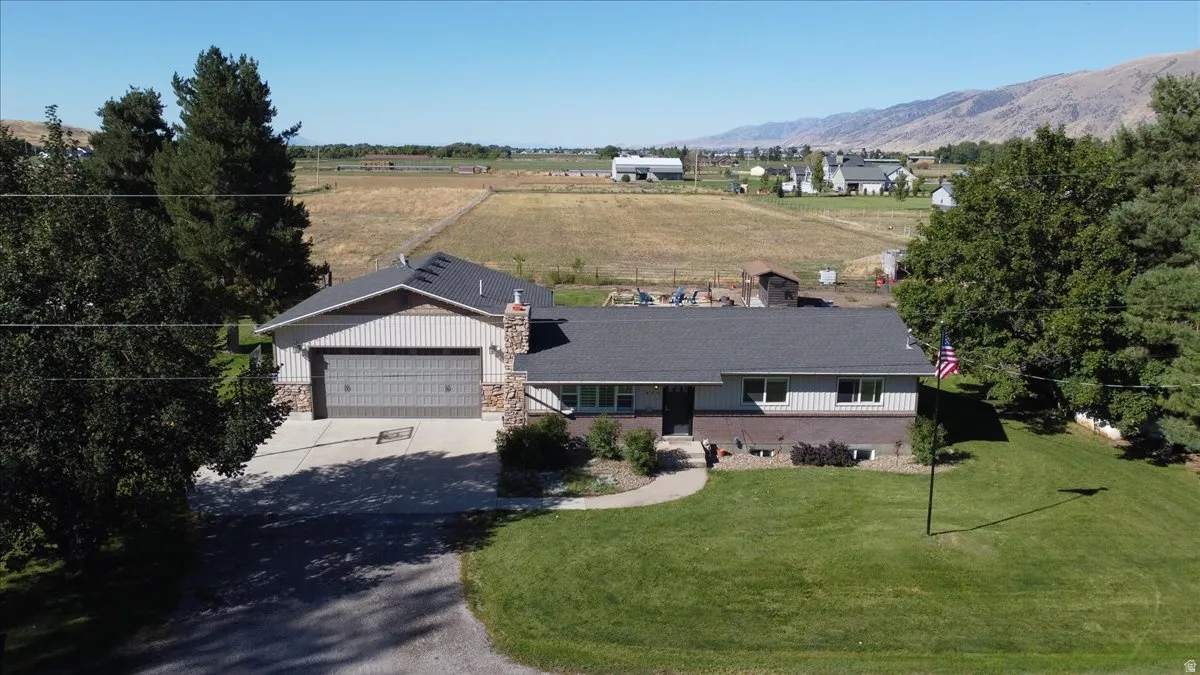 Expansive green lawn, mature trees, multi-level home with stone and siding exterior, attached garage, and mountain backdrop