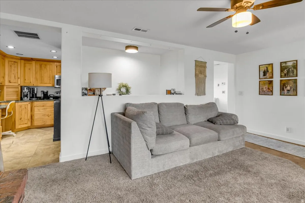 Interior living space featuring light neutral wall tones, a ceiling fan with integrated lighting, and a partial view into a kitchen with wood cabinetry and tile flooring