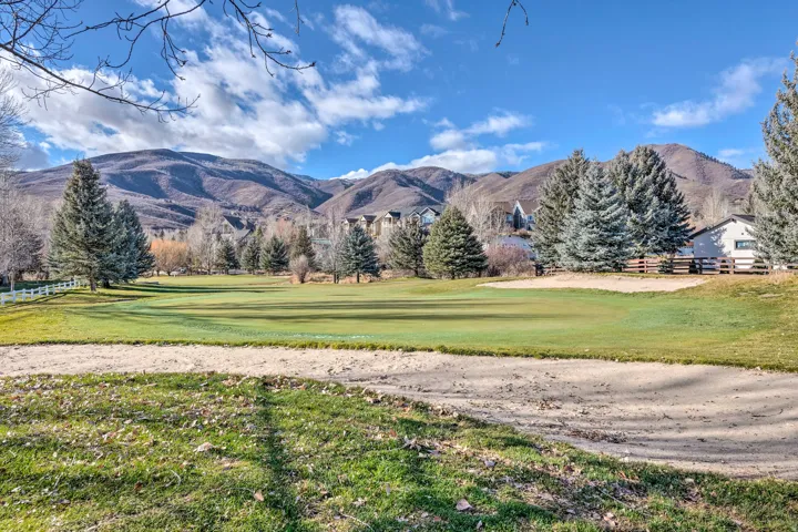 West view of expansive golf course green with sand traps, bordered by mature evergreen trees and a white split-rail fence
