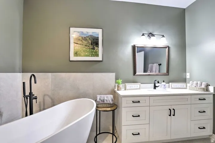 Freestanding soaking tub with a floor-mounted matte black faucet, wall-mounted light gray tiling, an extended white vanity with a single basin sink, and a framed wall mirror with overhead lighting