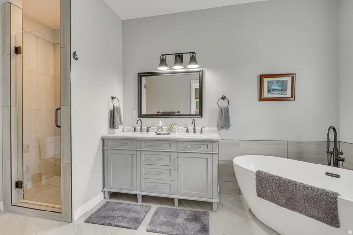 Freestanding soaking tub with a floor-mounted fixture, glass-enclosed shower, dual-sink vanity with a stone countertop, and wood-finish flooring