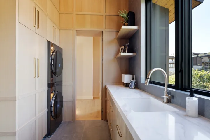 Integrated stacked laundry appliances, extensive light wood-finish cabinetry, white countertop with undermount sink, contemporary faucet, and floating shelves