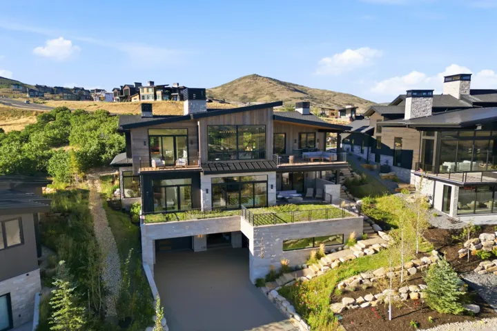 Multi-level architectural design featuring expansive glass walls, a covered drive-under garage, multiple outdoor terraces, and integrated landscaping with stone retaining walls