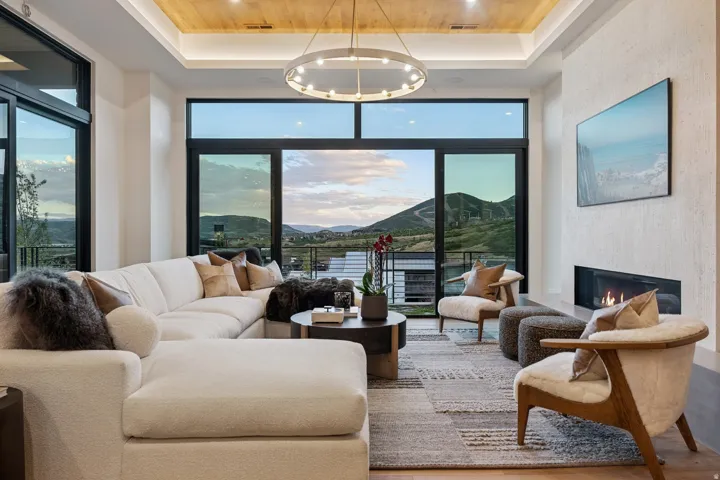 Expansive windows with dark frames, wood-paneled ceiling with recessed lighting, linear gas fireplace, and a contemporary chandelier