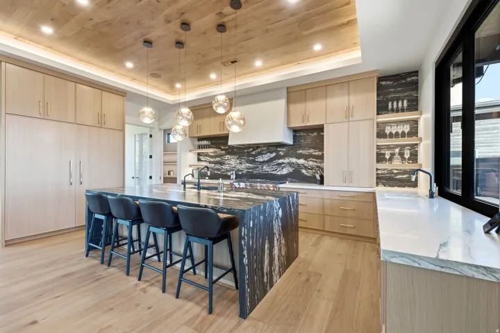 Kitchen featuring wood-finish flooring, custom wood-finish cabinetry, a large kitchen island with a stone countertop, a stone backsplash, and recessed and pendant lighting