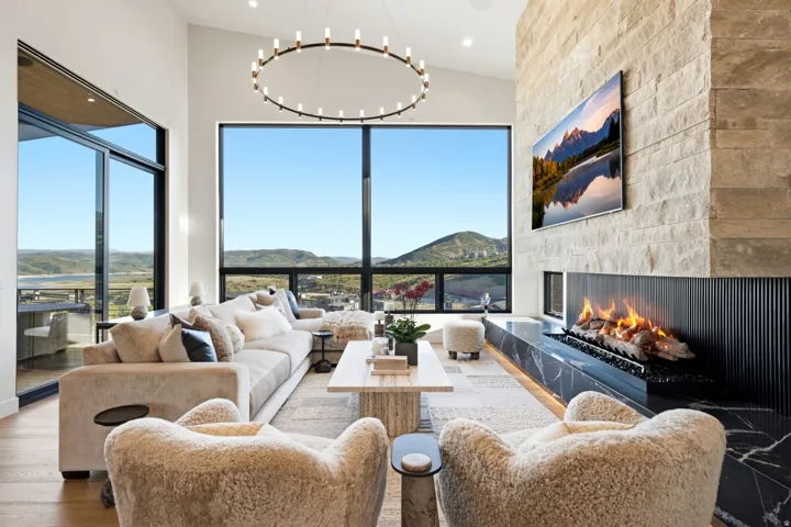Soaring ceilings with extensive window panels, stone-clad fireplace with a linear fire feature, wood-finish flooring, and a contemporary chandelier