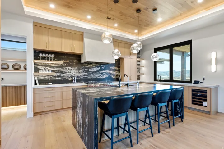Spacious kitchen featuring a large waterfall island, wood-finish flooring, dark stone backsplash, light wood cabinetry, and a built-in wine refrigerator