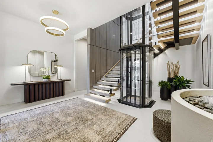 Open-tread staircase with integrated lighting, glass elevator, wood-finish wall paneling, and a contemporary chandelier