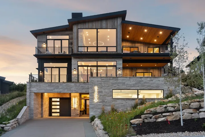Modern exterior showcasing multiple balconies, a stone-clad facade, large windows, an integrated garage, and a sloped, landscaped lot