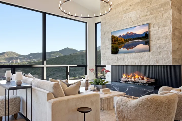 Expansive floor-to-ceiling windows showcasing mountain scenery, a stone fireplace with a black marble hearth, and a modern chandelier