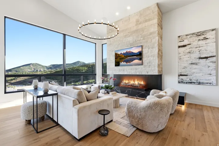 Expansive window wall, stone-clad fireplace with hearth, wide plank wood-finish flooring, and high ceilings with circular chandelier