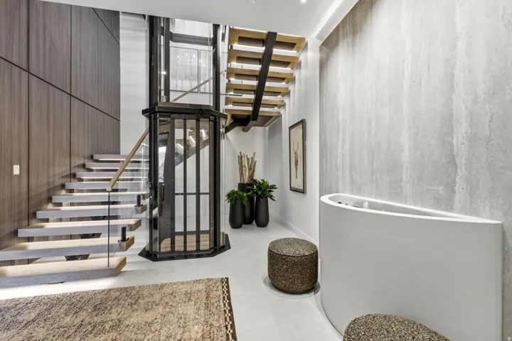 Modern glass elevator, open-tread staircase with glass railing, wood-paneled accent wall, and light gray wall finish