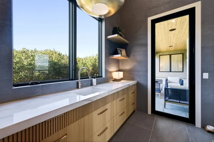 Integrated countertop sink, sleek black-framed window, textured wood-finish cabinetry, floating wood shelves, and a black-framed glass door