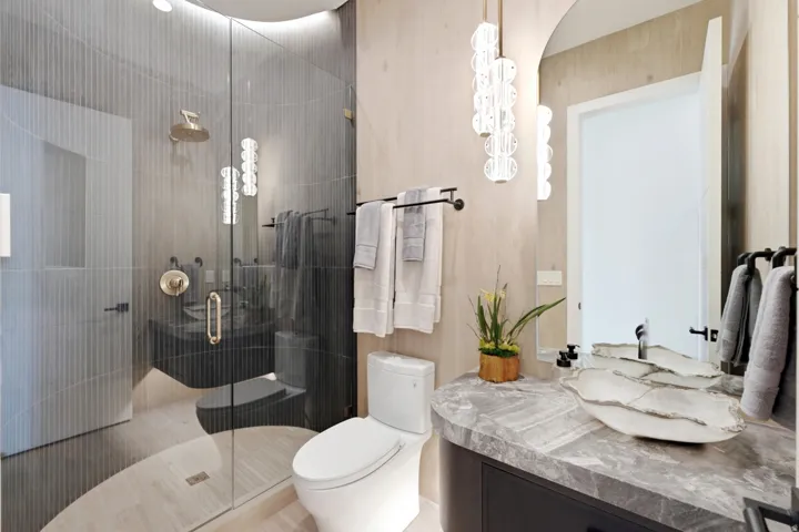Glass-enclosed shower with brass fixtures, a floating vanity with a vessel sink, a gray stone countertop, and contemporary pendant lighting