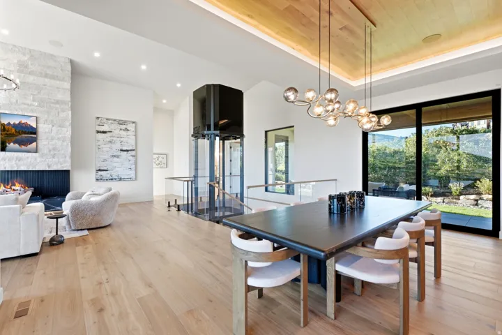 Light wood-finish flooring throughout, a glass-enclosed elevator, a stone-clad fireplace with a dark-finish hearth, a multi-globe chandelier, and expansive sliding glass doors