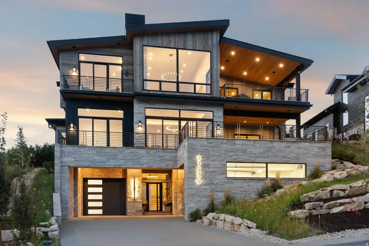 Modern architectural design with multiple balconies, a stacked stone and wood-paneled exterior, an integrated garage, and an exposed aggregate driveway