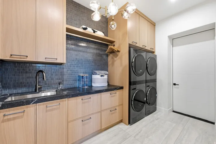 Laundry area featuring light wood-finish cabinetry, a dark countertop with a built-in sink, a blue tile backsplash, and a contemporary light fixture