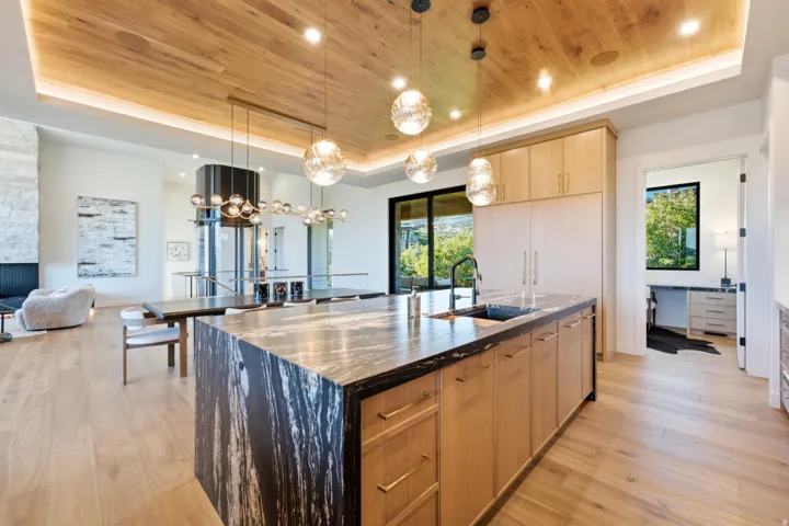 Kitchen island with a dark stone countertop and integrated sink, light wood-finish cabinetry with brass hardware, wood-finish flooring, and a wood-paneled ceiling with recessed and suspended lighting