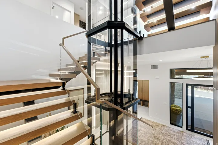 Central glass elevator, floating wood-finish staircase with glass railing, tall entryway doors, and a high ceiling with exposed wood beams