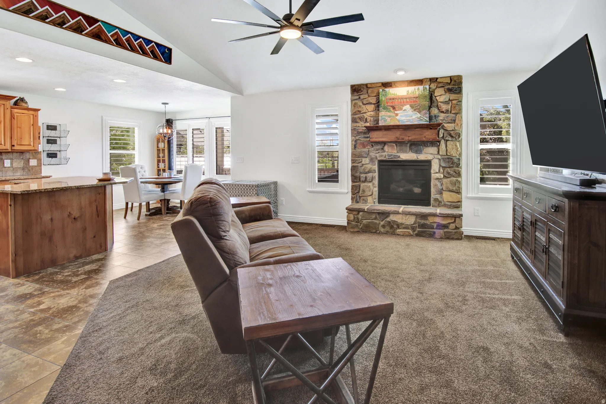 Vaulted ceiling with a ceiling fan, stone-faced fireplace with a mantel, plantation shutters, wood-finish kitchen cabinetry, and a built-in kitchen island with a stone countertop