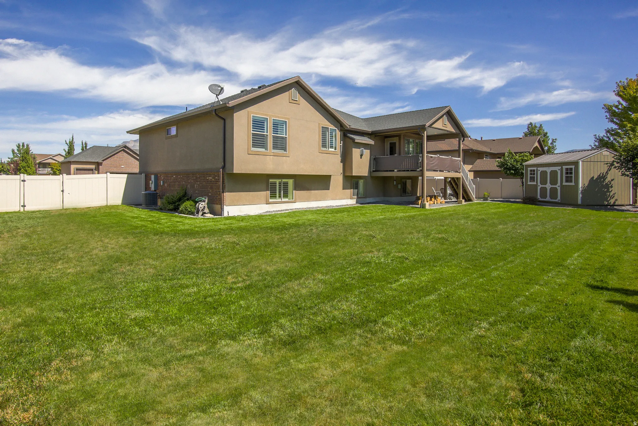 Expansive green lawn with a privacy fence, two-story exterior with stucco and brick accents, elevated deck with staircase, and an accessory storage shed
