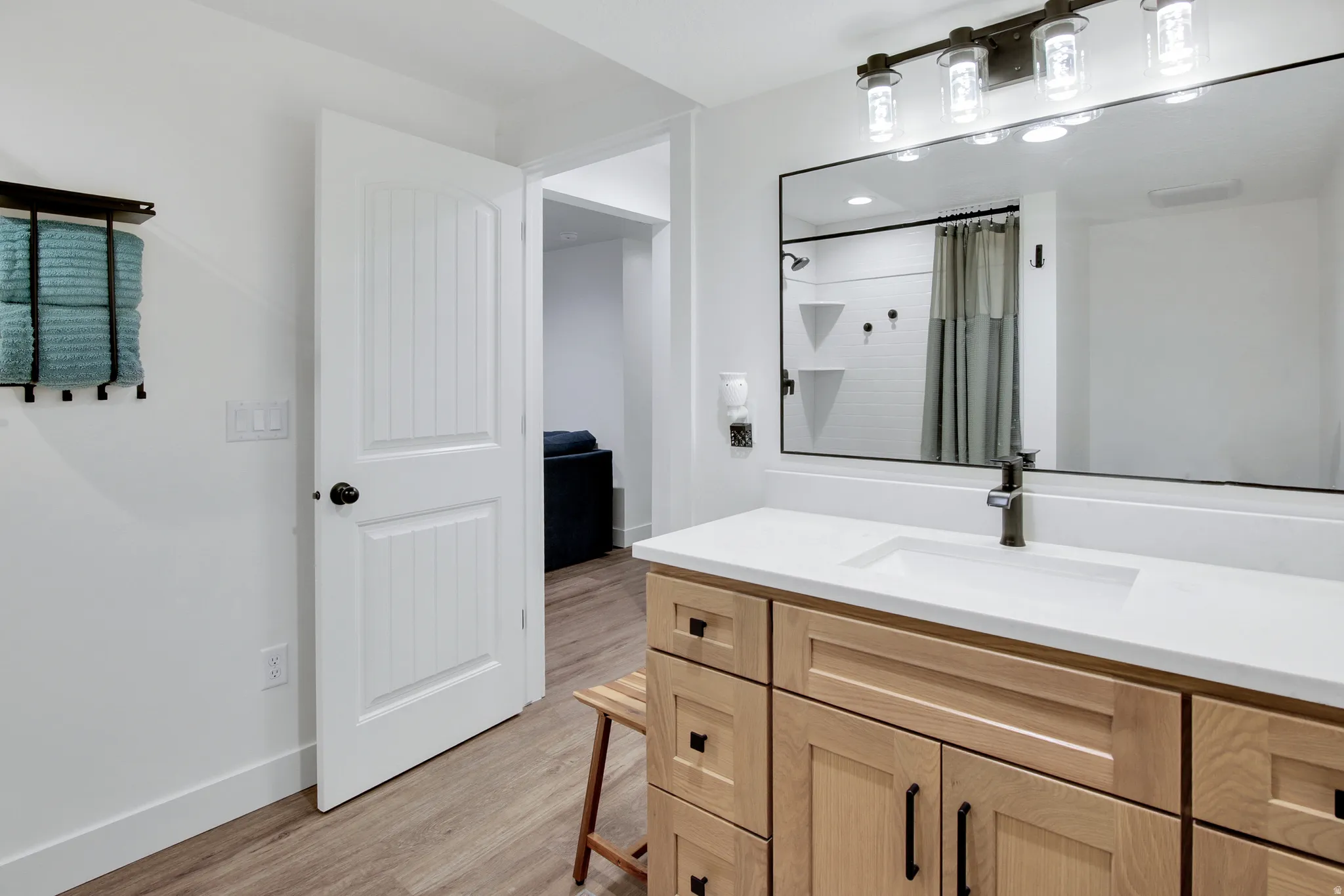 Wood-finish flooring, white paneled door, light-toned wood vanity with black hardware, white countertop with integrated sink, large rectangular mirror with black frame