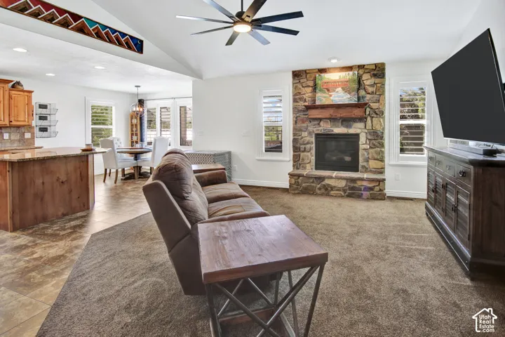 Vaulted ceiling with a ceiling fan, stone-faced fireplace with a mantel, plantation shutters, wood-finish kitchen cabinetry, and a built-in kitchen island with a stone countertop