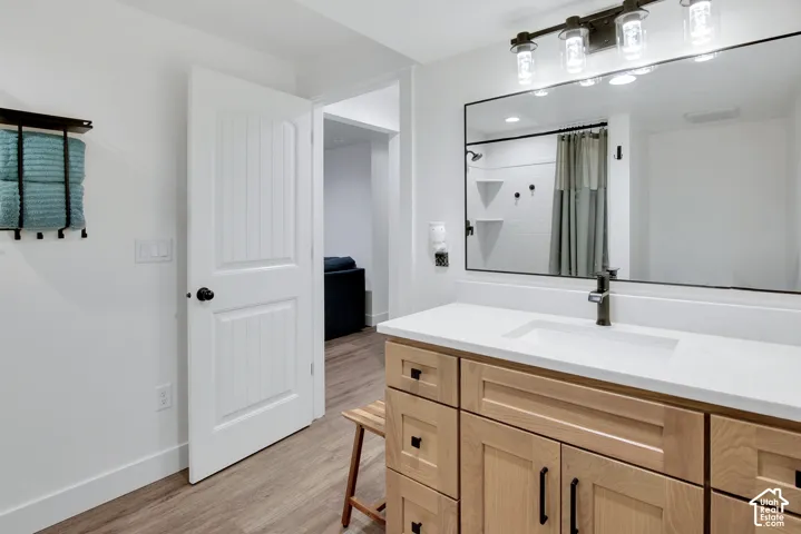 Wood-finish flooring, white paneled door, light-toned wood vanity with black hardware, white countertop with integrated sink, large rectangular mirror with black frame