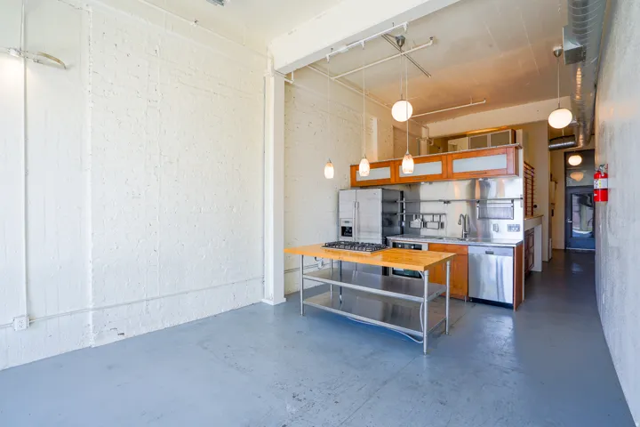 Open-concept space featuring a kitchen with stainless steel appliances, wood-finish cabinetry, and industrial pendant lighting