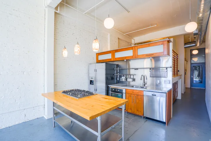 Kitchen featuring stainless steel appliances, wood-finish cabinetry, a freestanding butcher block island with integrated gas cooktop, and exposed brick walls